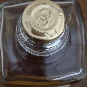 Pode incluir: Vista de cima de uma garrafa de vidro transparente com uma tampa dourada. A tampa apresenta um logotipo no centro. A garrafa cont&eacute;m um l&iacute;quido castanho. A imagem &eacute; tirada de uma perspectiva de cima.