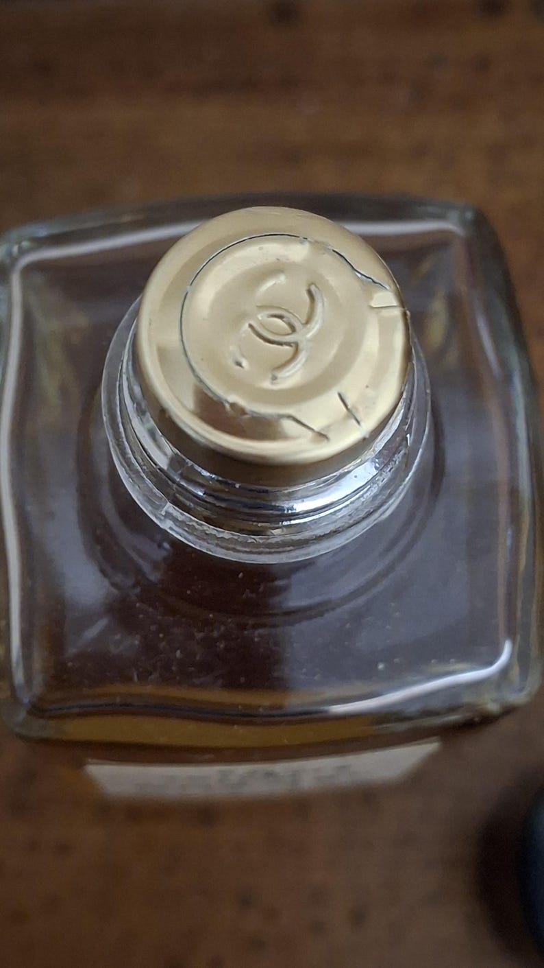 Pode incluir: Vista de cima de uma garrafa de vidro transparente com uma tampa dourada. A tampa apresenta um logotipo no centro. A garrafa cont&eacute;m um l&iacute;quido castanho. A imagem &eacute; tirada de uma perspectiva de cima.