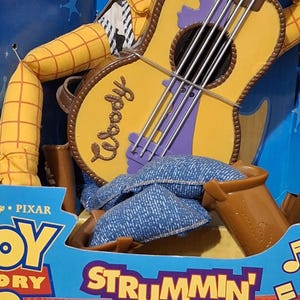 Puede incluir: Figura de juguete de Woody de Toy Story, sosteniendo una guitarra. La guitarra es amarilla con detalles morados y la palabra "Woody". El embalaje dice "Strummin' Singin' Woody".