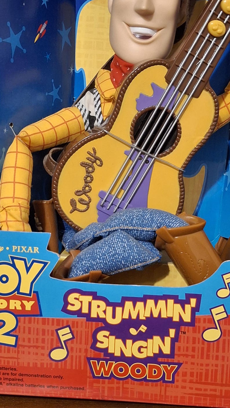 Puede incluir: Figura de juguete de Woody de Toy Story, sosteniendo una guitarra. La guitarra es amarilla con detalles morados y la palabra "Woody". El embalaje dice "Strummin' Singin' Woody".