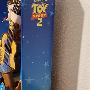 Puede incluir: Una figura de Woody en caja de Toy Story 2 de Disney Pixar. La caja es azul con gr&aacute;ficos de estrellas y el t&iacute;tulo de la pel&iacute;cula. Woody sostiene una guitarra. La parte inferior de la caja es roja.