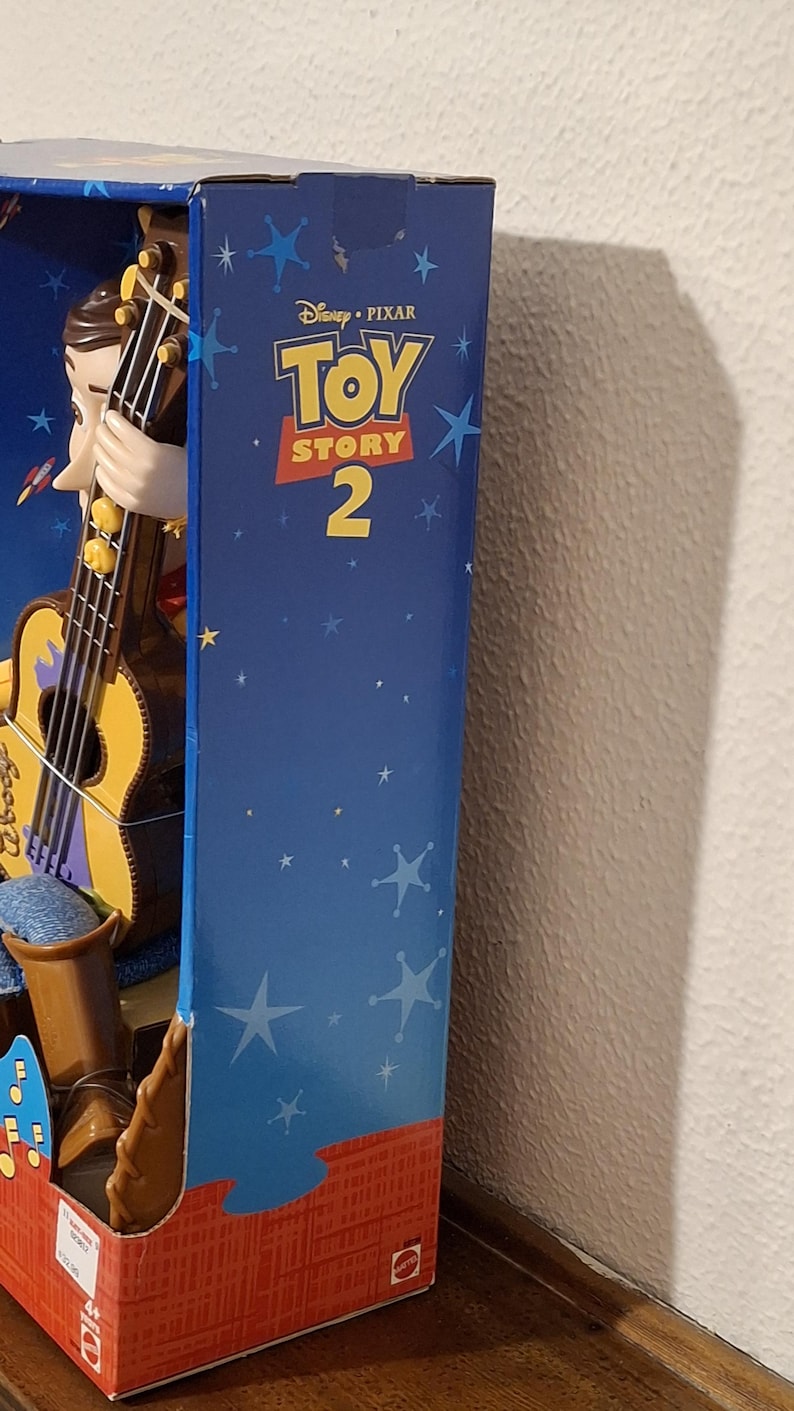 Puede incluir: Una figura de Woody en caja de Toy Story 2 de Disney Pixar. La caja es azul con gr&aacute;ficos de estrellas y el t&iacute;tulo de la pel&iacute;cula. Woody sostiene una guitarra. La parte inferior de la caja es roja.