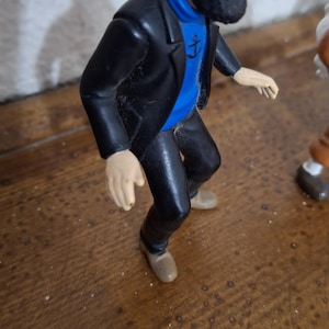K&ouml;nnte beinhalten: Eine Kunststofffigur eines b&auml;rtigen Charakters, der eine schwarze Kapit&auml;nsm&uuml;tze mit gelbem Band, eine schwarze Jacke, einen blauen Pullover mit schwarzem Ankeremblem und eine schwarze Hose tr&auml;gt. Die Figur ist in einer Vorw&auml;rtsneigung dargestellt.