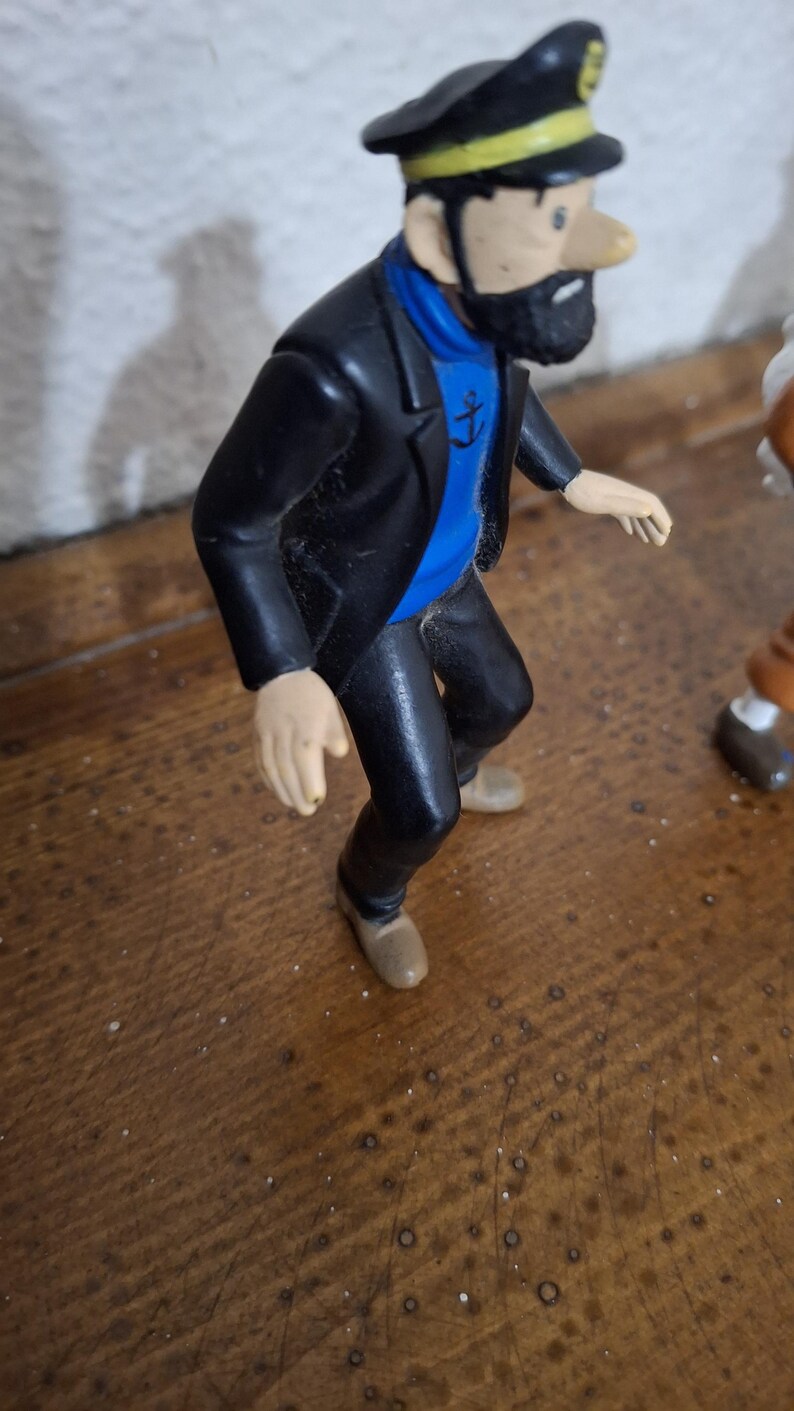 K&ouml;nnte beinhalten: Eine Kunststofffigur eines b&auml;rtigen Charakters, der eine schwarze Kapit&auml;nsm&uuml;tze mit gelbem Band, eine schwarze Jacke, einen blauen Pullover mit schwarzem Ankeremblem und eine schwarze Hose tr&auml;gt. Die Figur ist in einer Vorw&auml;rtsneigung dargestellt.
