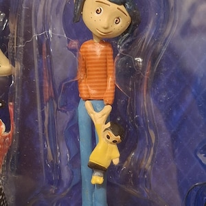 K&ouml;nnte beinhalten: Eine Figur einer Figur aus dem Animationsfilm "Coraline". Die Figur tr&auml;gt ein orange gestreiftes Hemd, eine blaue Hose und gelbe Schuhe. Die Figur h&auml;lt eine kleinere Figur in einem gelben Mantel.