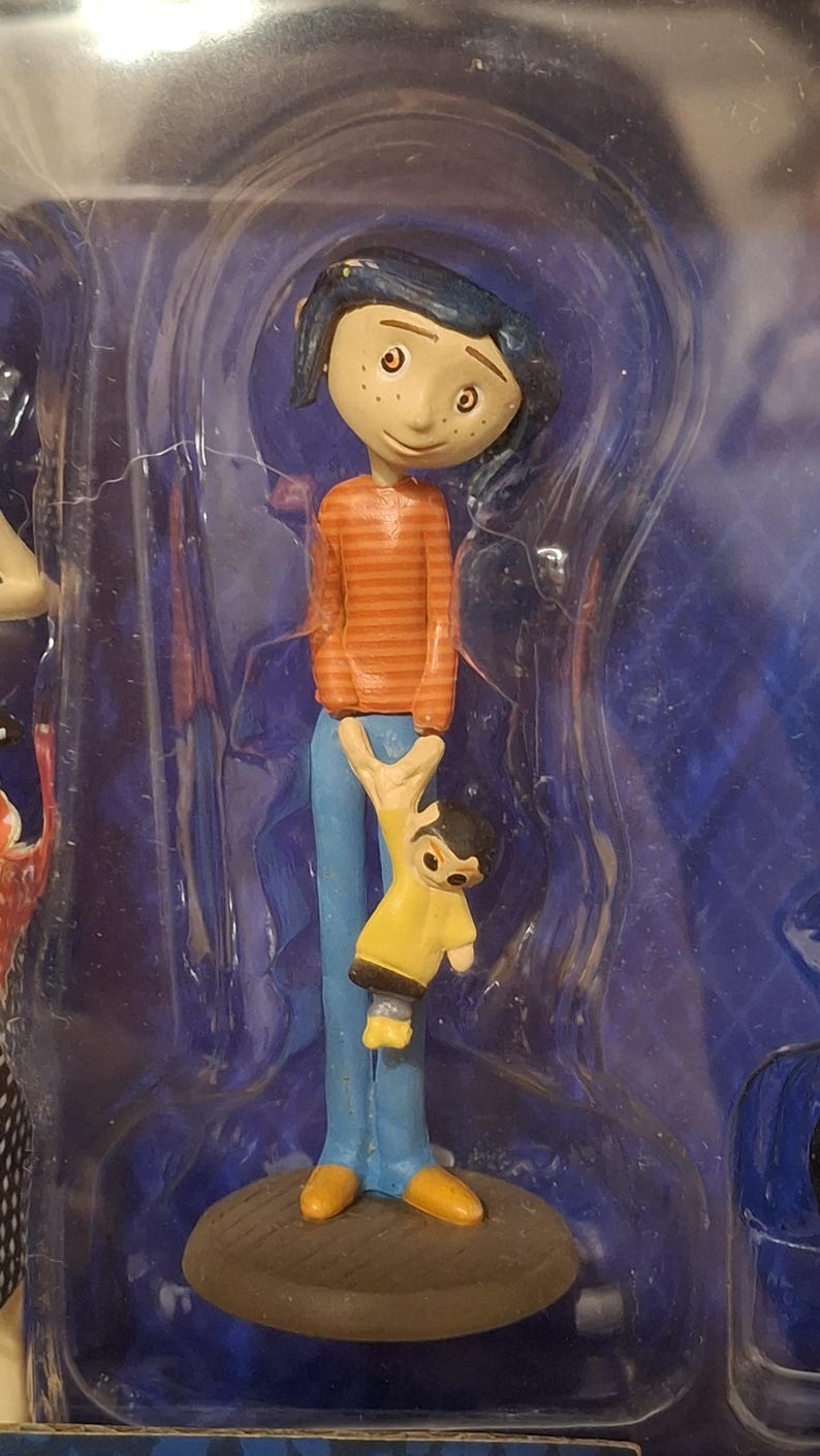 K&ouml;nnte beinhalten: Eine Figur einer Figur aus dem Animationsfilm "Coraline". Die Figur tr&auml;gt ein orange gestreiftes Hemd, eine blaue Hose und gelbe Schuhe. Die Figur h&auml;lt eine kleinere Figur in einem gelben Mantel.