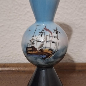 Puede incluir: Un jarrón decorativo de cerámica con la parte superior azul y la base negra, con detalles dorados. El centro esférico presenta una escena náutica pintada a mano de un velero en el océano. Un artículo de decoración para el hogar único.