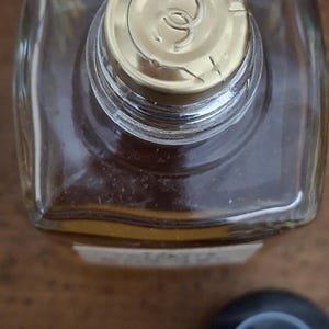 Pode incluir: Vista superior de um frasco de perfume transparente e quadrado com uma tampa dourada. A tampa apresenta um logotipo. Uma tampa preta separada com o mesmo logotipo est&aacute; ao lado do frasco. O frasco cont&eacute;m um l&iacute;quido castanho claro.