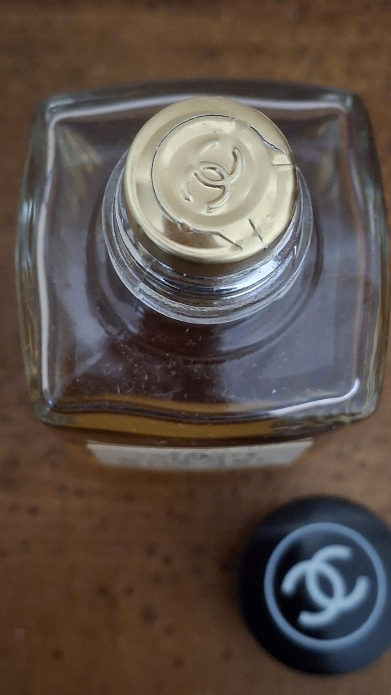 Pode incluir: Vista superior de um frasco de perfume transparente e quadrado com uma tampa dourada. A tampa apresenta um logotipo. Uma tampa preta separada com o mesmo logotipo est&aacute; ao lado do frasco. O frasco cont&eacute;m um l&iacute;quido castanho claro.