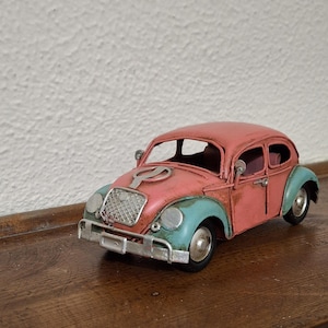 Puede incluir: Un coche de juguete de metal de estilo vintage con carrocería rosa, guardabarros verde azulado, parrilla delantera plateada, faros redondos y parachoques plateado. El modelo incluye un emblema circular en el capó y pequeños espejos laterales.