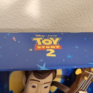 Puede incluir: Caja azul con el logo "Toy Story 2", mostrando la cara de Woody y su guitarra. La caja est&aacute; decorada con un dise&ntilde;o de cielo estrellado, cohetes y estrellas. Tambi&eacute;n se ve el logo de Disney Pixar.
