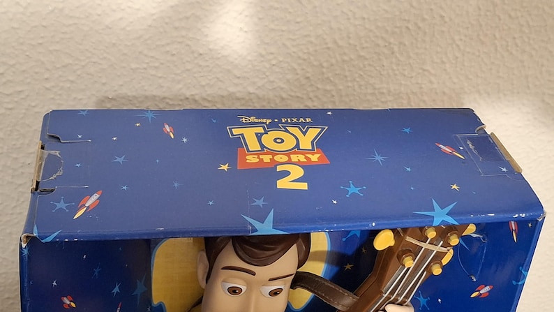 Puede incluir: Caja azul con el logo "Toy Story 2", mostrando la cara de Woody y su guitarra. La caja est&aacute; decorada con un dise&ntilde;o de cielo estrellado, cohetes y estrellas. Tambi&eacute;n se ve el logo de Disney Pixar.