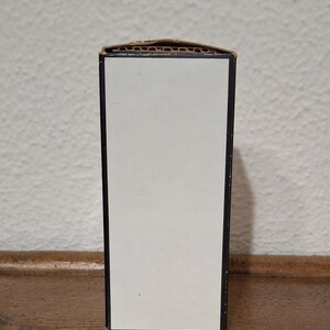 Pode incluir: Uma caixa retangular com um painel frontal branco e acabamento preto. A borda superior da caixa &eacute; feita de papel&atilde;o ondulado. A caixa est&aacute; colocada contra uma parede branca texturizada e uma superf&iacute;cie de madeira.