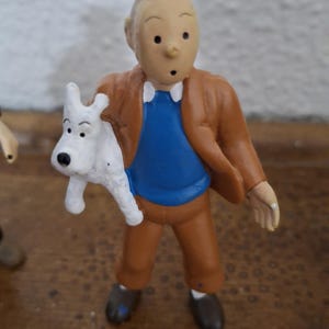 K&ouml;nnte beinhalten: Eine kleine Spielfigur einer Comicfigur mit charakteristischer Tolle, bekleidet mit einer braunen Jacke, einem blauen Pullover und einer braunen Hose, die einen kleinen wei&szlig;en Hund mit schwarzen Ohren und Nase im rechten Arm h&auml;lt.