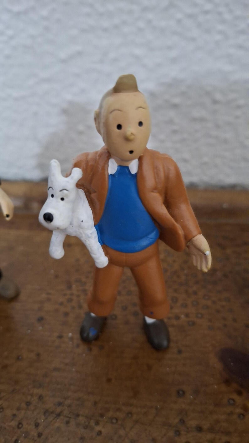 K&ouml;nnte beinhalten: Eine kleine Spielfigur einer Comicfigur mit charakteristischer Tolle, bekleidet mit einer braunen Jacke, einem blauen Pullover und einer braunen Hose, die einen kleinen wei&szlig;en Hund mit schwarzen Ohren und Nase im rechten Arm h&auml;lt.