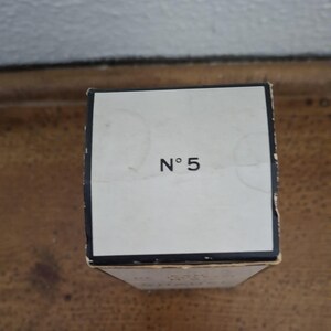 Pode incluir: Uma caixa retangular esbranqui&ccedil;ada com o texto "N&deg; 5" em preto. A caixa tem uma borda preta e est&aacute; colocada sobre uma superf&iacute;cie de madeira. O fundo &eacute; uma parede de cor clara.