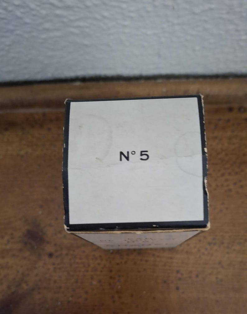 Pode incluir: Uma caixa retangular esbranqui&ccedil;ada com o texto "N&deg; 5" em preto. A caixa tem uma borda preta e est&aacute; colocada sobre uma superf&iacute;cie de madeira. O fundo &eacute; uma parede de cor clara.