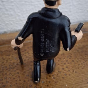 K&ouml;nnte beinhalten: Eine kleine Figur einer Person in einem schwarzen Anzug und mit schwarzer Melone, die einen d&uuml;nnen schwarzen Gehstock in einer Hand h&auml;lt. Auf dem R&uuml;cken der Jacke befindet sich der gepr&auml;gte Text HERG&Eacute; 1954 TL PLASTO und ein CE-Zeichen.
