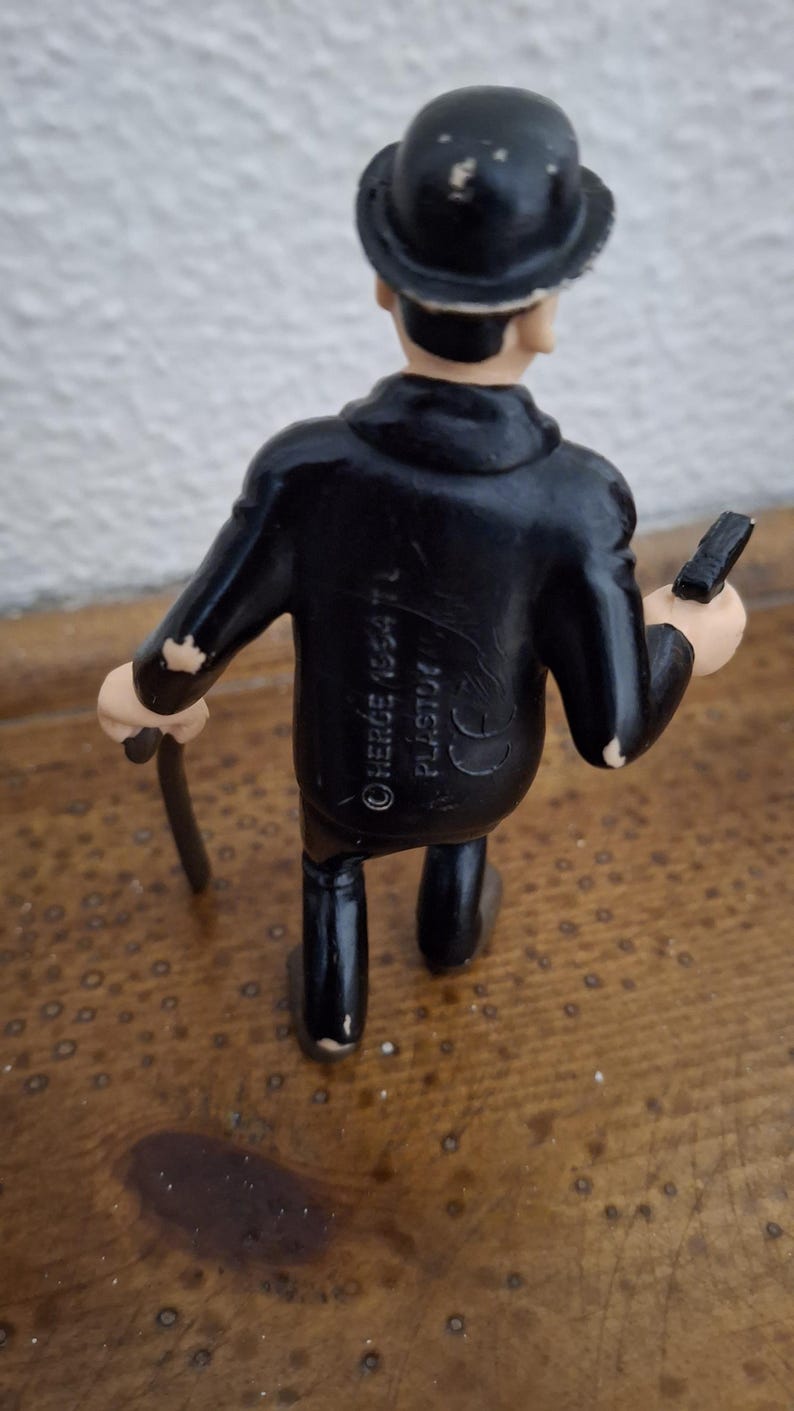 K&ouml;nnte beinhalten: Eine kleine Figur einer Person in einem schwarzen Anzug und mit schwarzer Melone, die einen d&uuml;nnen schwarzen Gehstock in einer Hand h&auml;lt. Auf dem R&uuml;cken der Jacke befindet sich der gepr&auml;gte Text HERG&Eacute; 1954 TL PLASTO und ein CE-Zeichen.
