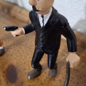 K&ouml;nnte beinhalten: Eine kleine Figur einer Person in einem schwarzen Anzug, einem wei&szlig;en Hemd mit schwarzer Krawatte und einer schwarzen Melone. Die Figur hat einen markanten Schnurrbart, h&auml;lt eine kleine schwarze Pistole in der rechten und einen d&uuml;nnen braunen Stock in der linken Hand.
