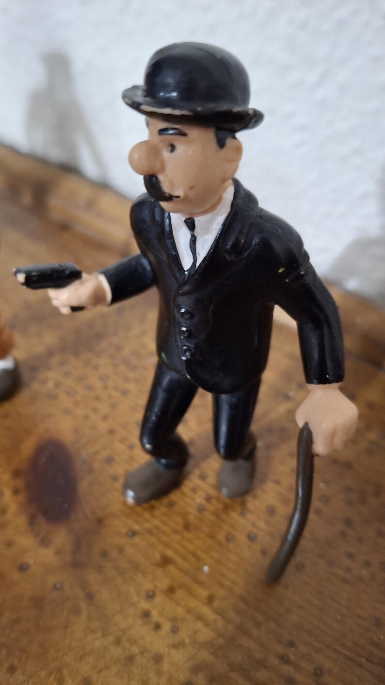 K&ouml;nnte beinhalten: Eine kleine Figur einer Person in einem schwarzen Anzug, einem wei&szlig;en Hemd mit schwarzer Krawatte und einer schwarzen Melone. Die Figur hat einen markanten Schnurrbart, h&auml;lt eine kleine schwarze Pistole in der rechten und einen d&uuml;nnen braunen Stock in der linken Hand.
