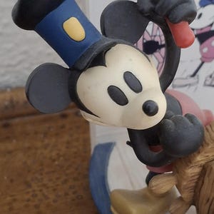 May include: A Mickey Mouse figurine in a Steamboat Willie theme. Mickey is wearing a blue hat with a yellow accent and black gloves. The figurine is on a boat with a blue and white design.