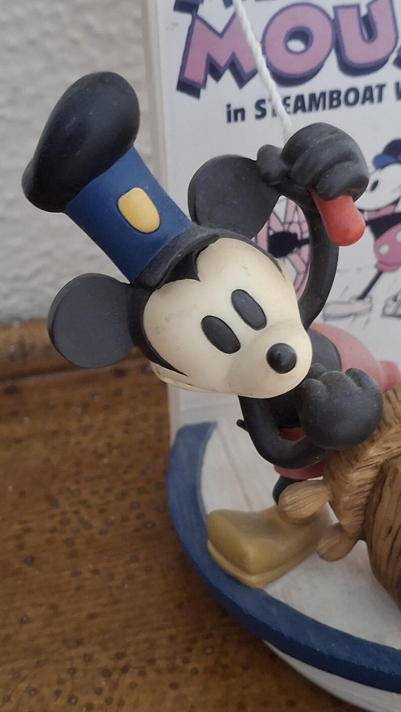 May include: A Mickey Mouse figurine in a Steamboat Willie theme. Mickey is wearing a blue hat with a yellow accent and black gloves. The figurine is on a boat with a blue and white design.
