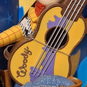 Puede incluir: Figura de Woody con una guitarra amarilla. La guitarra tiene bordes marrones, detalles morados y el nombre "Woody". La figura lleva un pa&ntilde;uelo rojo y una camisa amarilla.
