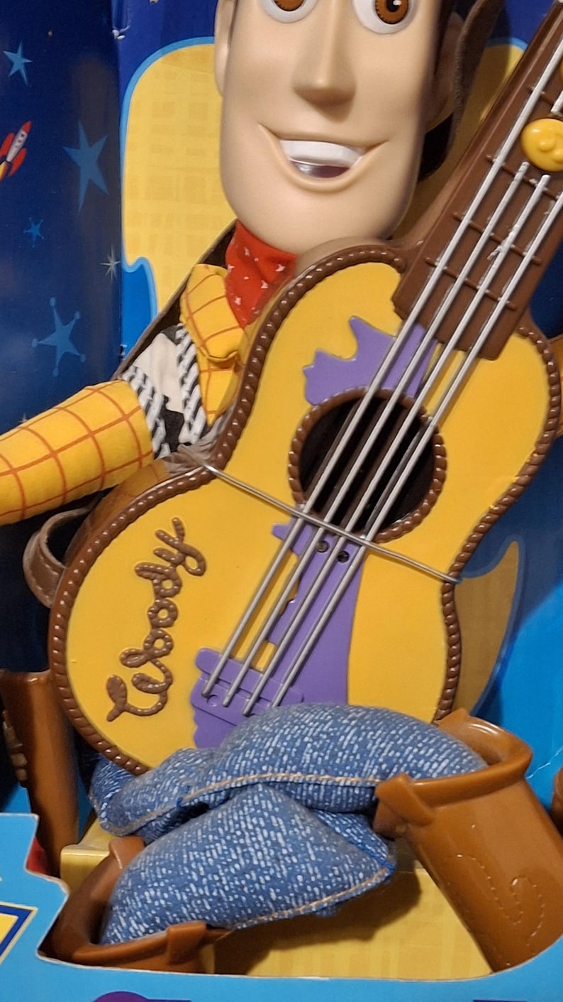 Puede incluir: Figura de Woody con una guitarra amarilla. La guitarra tiene bordes marrones, detalles morados y el nombre "Woody". La figura lleva un pa&ntilde;uelo rojo y una camisa amarilla.