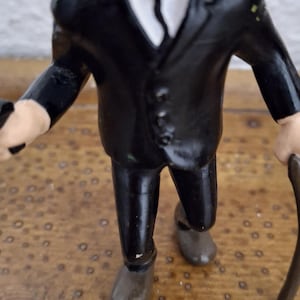 K&ouml;nnte beinhalten: Eine kleine Figur einer Person in einem schwarzen Anzug mit wei&szlig;em Hemd und schwarzer Krawatte. Die Figur hat einen markanten schwarzen Schnurrbart, helle Haut und h&auml;lt einen dunklen Gehstock in einer Hand, w&auml;hrend sie aufrecht steht.