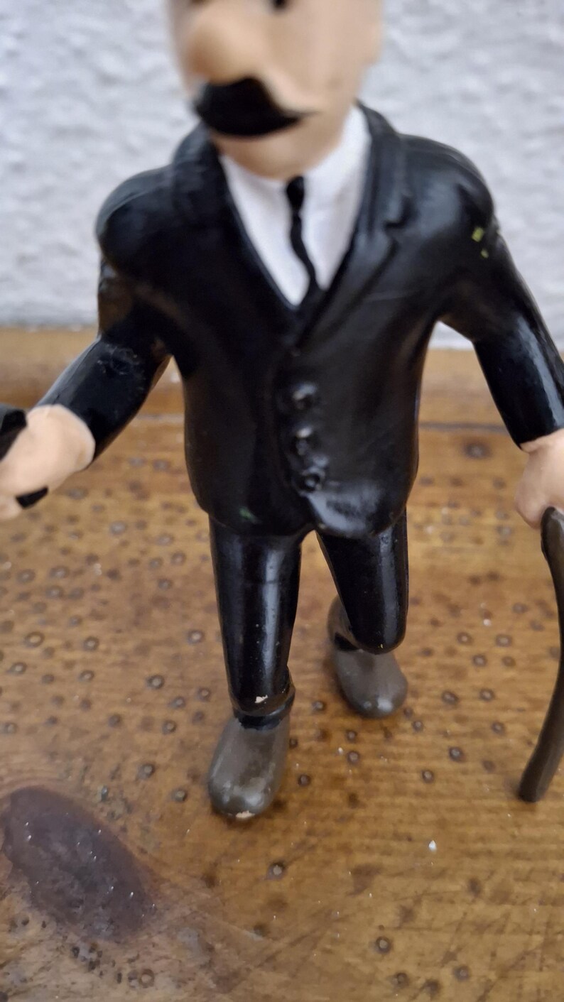 K&ouml;nnte beinhalten: Eine kleine Figur einer Person in einem schwarzen Anzug mit wei&szlig;em Hemd und schwarzer Krawatte. Die Figur hat einen markanten schwarzen Schnurrbart, helle Haut und h&auml;lt einen dunklen Gehstock in einer Hand, w&auml;hrend sie aufrecht steht.