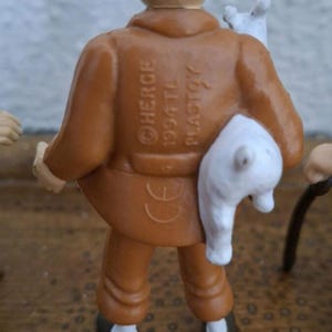 K&ouml;nnte beinhalten: Eine kleine Figur einer Person in brauner Jacke und Hose, die einen wei&szlig;en Hund h&auml;lt. Auf dem R&uuml;cken der Jacke befindet sich der gepr&auml;gte Text HERGE 1994 TL PLASTOY sowie ein CE-Zeichen. Die Figur hat helle Haut und dunkle Schuhe.