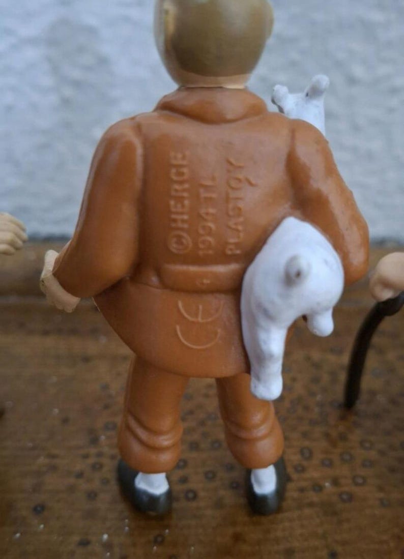K&ouml;nnte beinhalten: Eine kleine Figur einer Person in brauner Jacke und Hose, die einen wei&szlig;en Hund h&auml;lt. Auf dem R&uuml;cken der Jacke befindet sich der gepr&auml;gte Text HERGE 1994 TL PLASTOY sowie ein CE-Zeichen. Die Figur hat helle Haut und dunkle Schuhe.