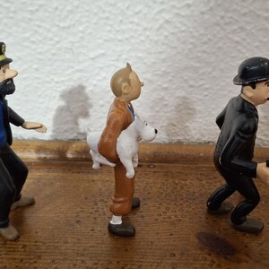 K&ouml;nnte beinhalten: Drei Kunststofffiguren in einer Reihe. Die erste ist ein b&auml;rtiger Mann mit schwarzer Kapit&auml;nsm&uuml;tze, blauem Pullover und schwarzer Hose. Die zweite ist ein Mann in brauner Jacke und Hose, der einen wei&szlig;en Hund h&auml;lt. Die dritte ist ein Mann im schwarzen Anzug mit Melone, der einen kleinen schwarzen Gegenstand h&auml;lt.