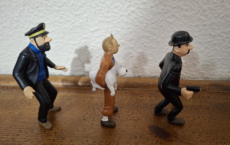 K&ouml;nnte beinhalten: Drei Kunststofffiguren in einer Reihe. Die erste ist ein b&auml;rtiger Mann mit schwarzer Kapit&auml;nsm&uuml;tze, blauem Pullover und schwarzer Hose. Die zweite ist ein Mann in brauner Jacke und Hose, der einen wei&szlig;en Hund h&auml;lt. Die dritte ist ein Mann im schwarzen Anzug mit Melone, der einen kleinen schwarzen Gegenstand h&auml;lt.