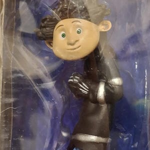 K&ouml;nnte beinhalten: Figur einer Figur mit brauner Haut, gr&uuml;nen Augen und dunklem, lockigem Haar. Die Figur tr&auml;gt einen schwarzen Mantel mit silbernen Verzierungen, eine blaue Hose und dunkelgr&uuml;ne Schuhe. Die Figur steht auf einem Steinsockel.