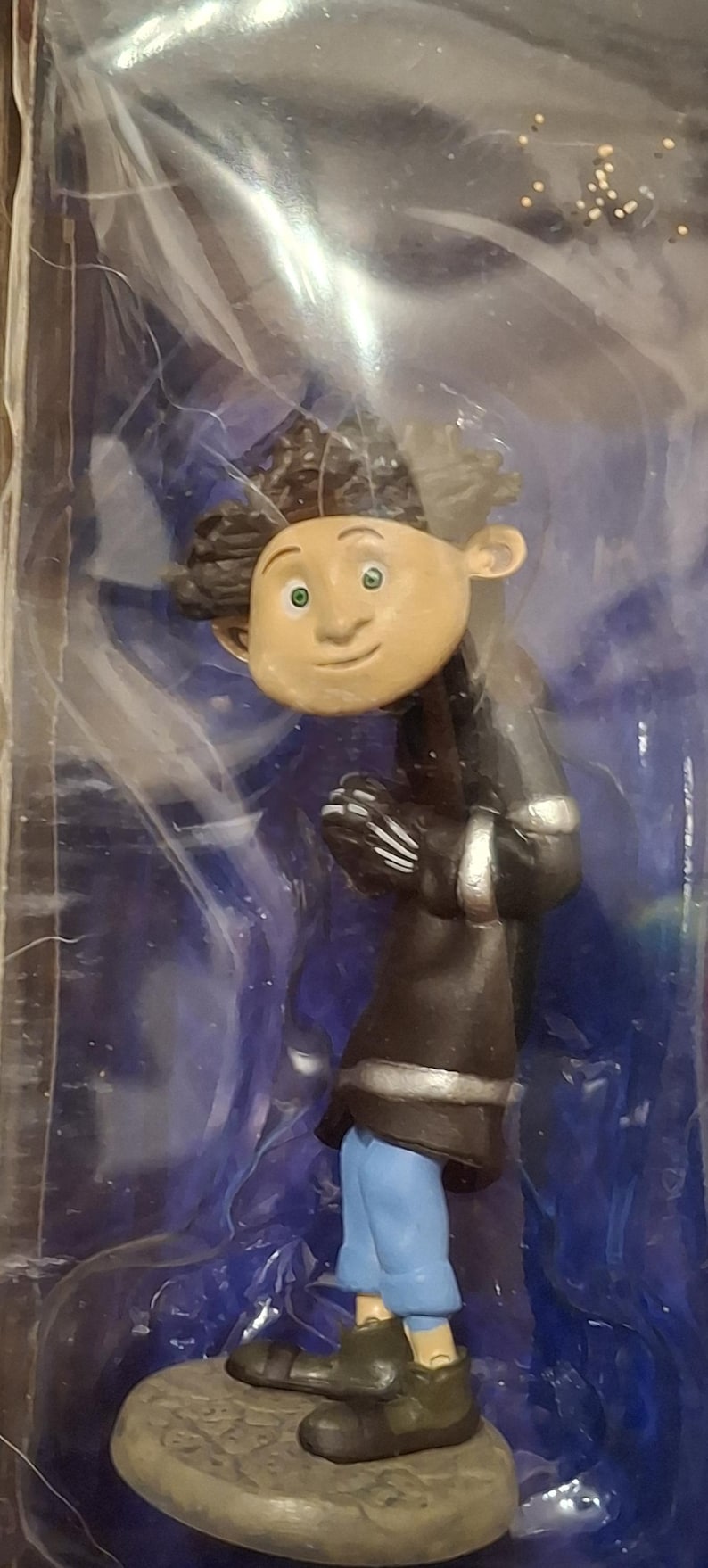 K&ouml;nnte beinhalten: Figur einer Figur mit brauner Haut, gr&uuml;nen Augen und dunklem, lockigem Haar. Die Figur tr&auml;gt einen schwarzen Mantel mit silbernen Verzierungen, eine blaue Hose und dunkelgr&uuml;ne Schuhe. Die Figur steht auf einem Steinsockel.