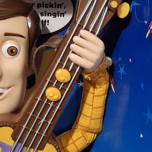 Puede incluir: Figura de juguete de Woody sosteniendo una guitarra marr&oacute;n y amarilla. La guitarra tiene cuerdas plateadas y clavijas de afinaci&oacute;n amarillas. La figura lleva una camisa a cuadros amarilla. Fondo azul con estrellas blancas.