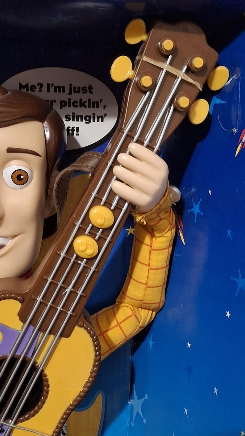 Puede incluir: Figura de juguete de Woody sosteniendo una guitarra marr&oacute;n y amarilla. La guitarra tiene cuerdas plateadas y clavijas de afinaci&oacute;n amarillas. La figura lleva una camisa a cuadros amarilla. Fondo azul con estrellas blancas.