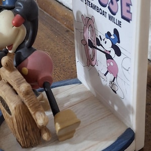 May include: A Mickey Mouse figurine from Steamboat Willie. The figurine features Mickey Mouse in his classic outfit, with a black body, red shorts, and yellow shoes. The base is white with a blue trim. The text "Mickey Mouse in Steamboat Willie" is visible.