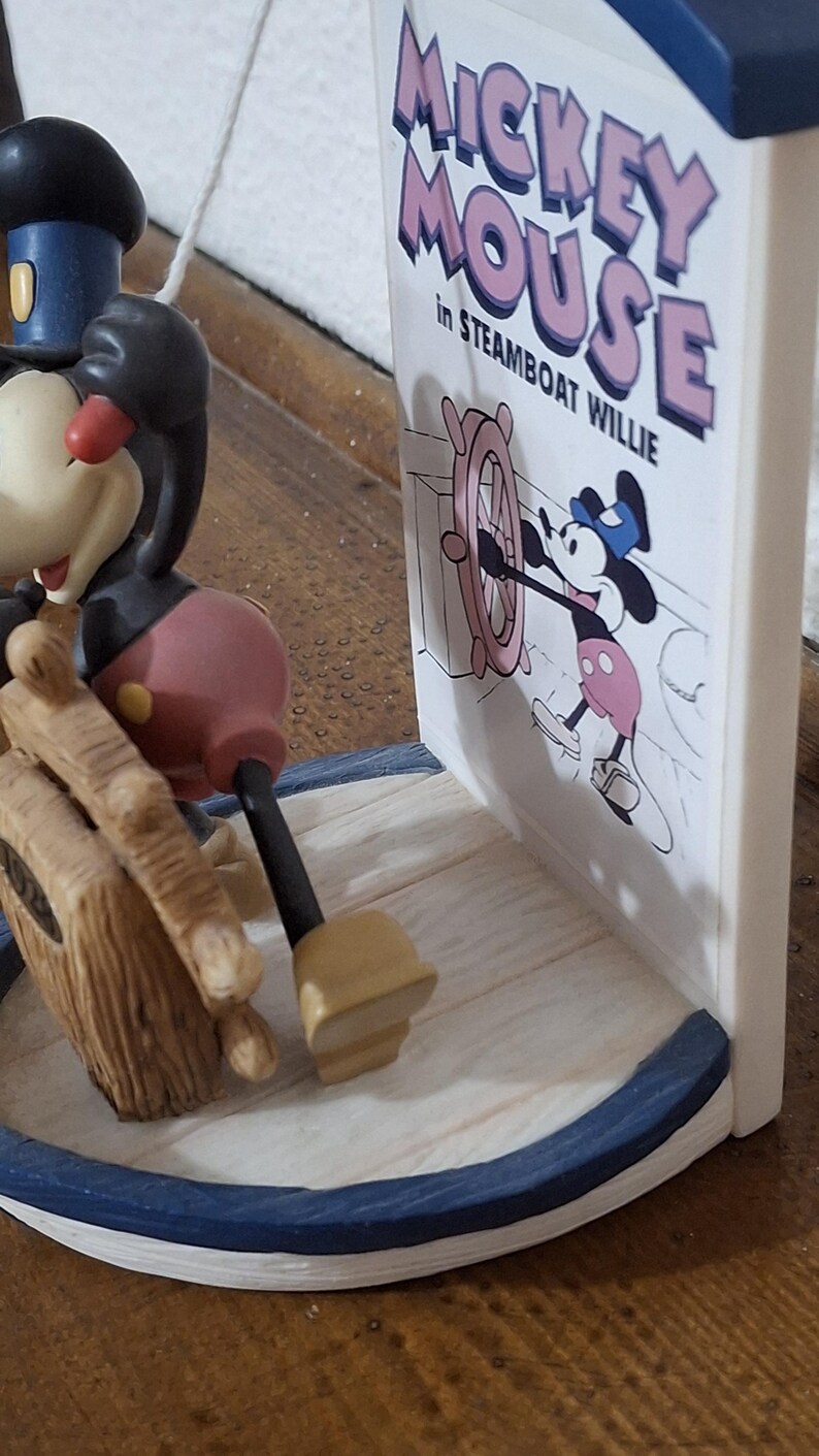 May include: A Mickey Mouse figurine from Steamboat Willie. The figurine features Mickey Mouse in his classic outfit, with a black body, red shorts, and yellow shoes. The base is white with a blue trim. The text "Mickey Mouse in Steamboat Willie" is visible.