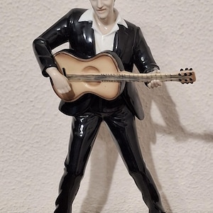 May include: A ceramic figurine of a man in a black suit and white shirt, playing a guitar. The figurine is on a white base and has a glossy finish. The guitar is tan and brown.