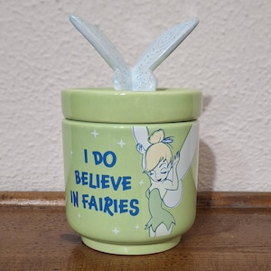 Può includere: Un barattolo in ceramica verde chiaro con coperchio e manico a forma di farfalla. Il barattolo presenta la scritta "I DO BELIEVE IN FAIRIES" e un'illustrazione di Trilli.