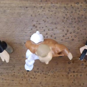 K&ouml;nnte beinhalten: Drei kleine Figuren von oben betrachtet. Die linke Figur tr&auml;gt einen schwarzen Anzug und eine M&uuml;tze mit gr&uuml;nem Streifen. Die mittlere Figur ist hellbraun und tr&auml;gt wei&szlig;e Kleidung. Die rechte Figur tr&auml;gt einen schwarzen Anzug und Hut mit Objekt.