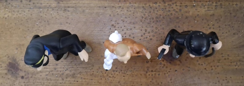 K&ouml;nnte beinhalten: Drei kleine Figuren von oben betrachtet. Die linke Figur tr&auml;gt einen schwarzen Anzug und eine M&uuml;tze mit gr&uuml;nem Streifen. Die mittlere Figur ist hellbraun und tr&auml;gt wei&szlig;e Kleidung. Die rechte Figur tr&auml;gt einen schwarzen Anzug und Hut mit Objekt.