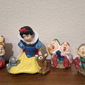 Pode incluir: Um conjunto de quatro figuras de cerâmica com a Branca de Neve num vestido azul e amarelo com um coelho e um veado, ao lado de três grupos dos sete anões, incluindo Grumpy, Happy, Sleepy, Sneezy, Bashful, Doc e Dopey, com nomes inscritos.