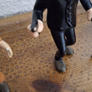 K&ouml;nnte beinhalten: Eine kleine Figur einer Person in einem schwarzen Anzug mit wei&szlig;em Hemd und schwarzer Krawatte, die einen markanten schwarzen Schnurrbart tr&auml;gt. Die Figur h&auml;lt einen kleinen schwarzen Gegenstand in einer Hand und tr&auml;gt braune Schuhe.