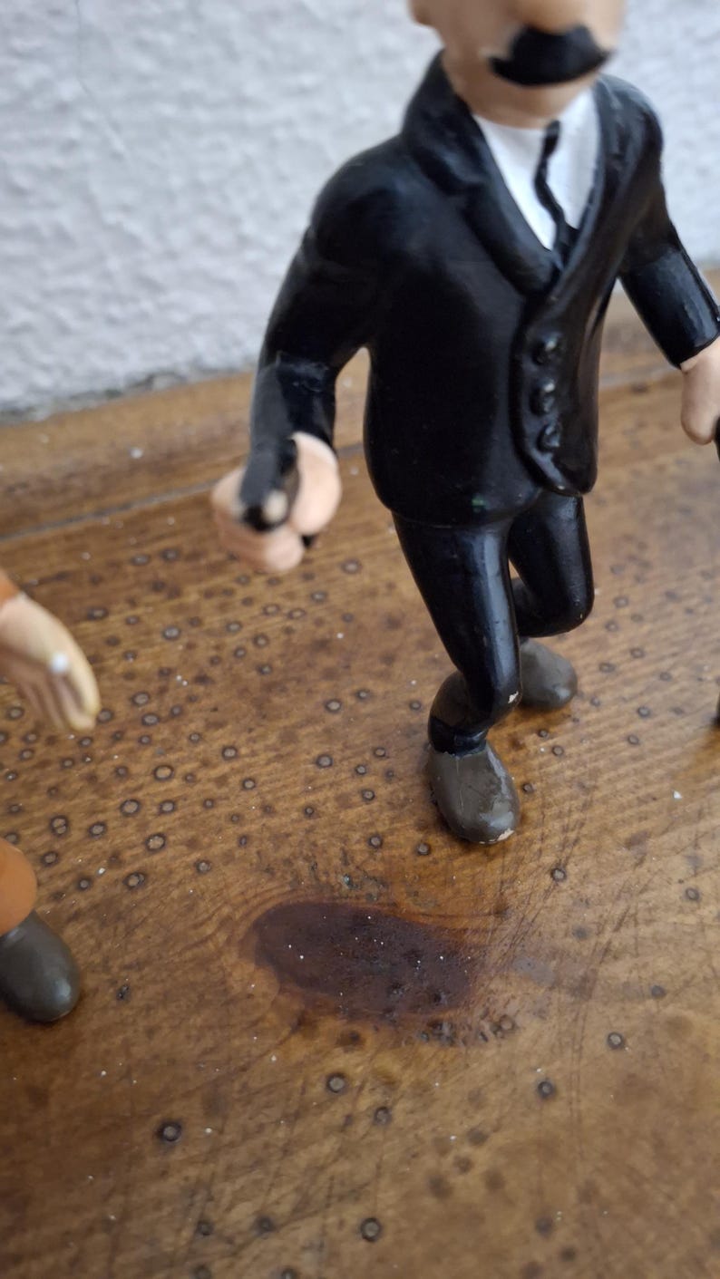 K&ouml;nnte beinhalten: Eine kleine Figur einer Person in einem schwarzen Anzug mit wei&szlig;em Hemd und schwarzer Krawatte, die einen markanten schwarzen Schnurrbart tr&auml;gt. Die Figur h&auml;lt einen kleinen schwarzen Gegenstand in einer Hand und tr&auml;gt braune Schuhe.