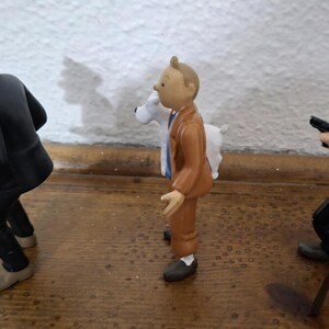 K&ouml;nnte beinhalten: Ein Set aus drei Kunststofffiguren, bestehend aus einem Mann in schwarzem Anzug und M&uuml;tze, einem Mann in braunem Anzug mit einem wei&szlig;en Hund und einem Mann in schwarzem Anzug mit Melone, der einen kleinen schwarzen Gegenstand h&auml;lt, auf einer Holzfl&auml;che.