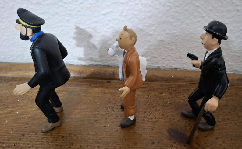 K&ouml;nnte beinhalten: Ein Set aus drei Kunststofffiguren, bestehend aus einem Mann in schwarzem Anzug und M&uuml;tze, einem Mann in braunem Anzug mit einem wei&szlig;en Hund und einem Mann in schwarzem Anzug mit Melone, der einen kleinen schwarzen Gegenstand h&auml;lt, auf einer Holzfl&auml;che.